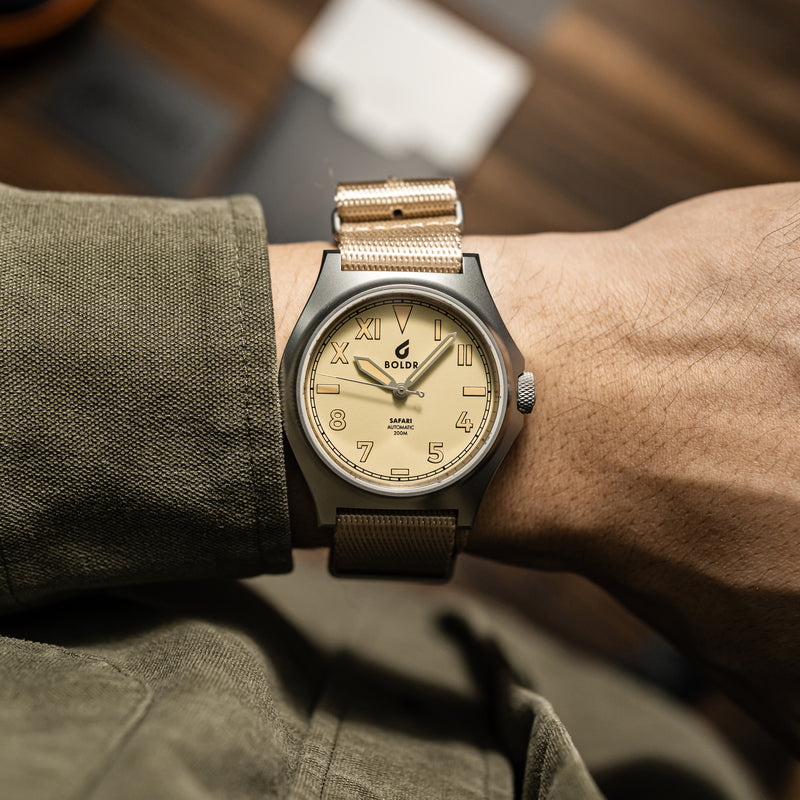 Close-up of a BOLDR watch with a beige dial and matching beige fabric strap, placed on a wooden surface. The watch face displays a mix of Roman and Arabic numerals, with the brand name ‘BOLDR’ and ‘Safari Automatic 200M’ text prominently visible. The watch is neatly arranged, showcasing its design and craftsmanship.