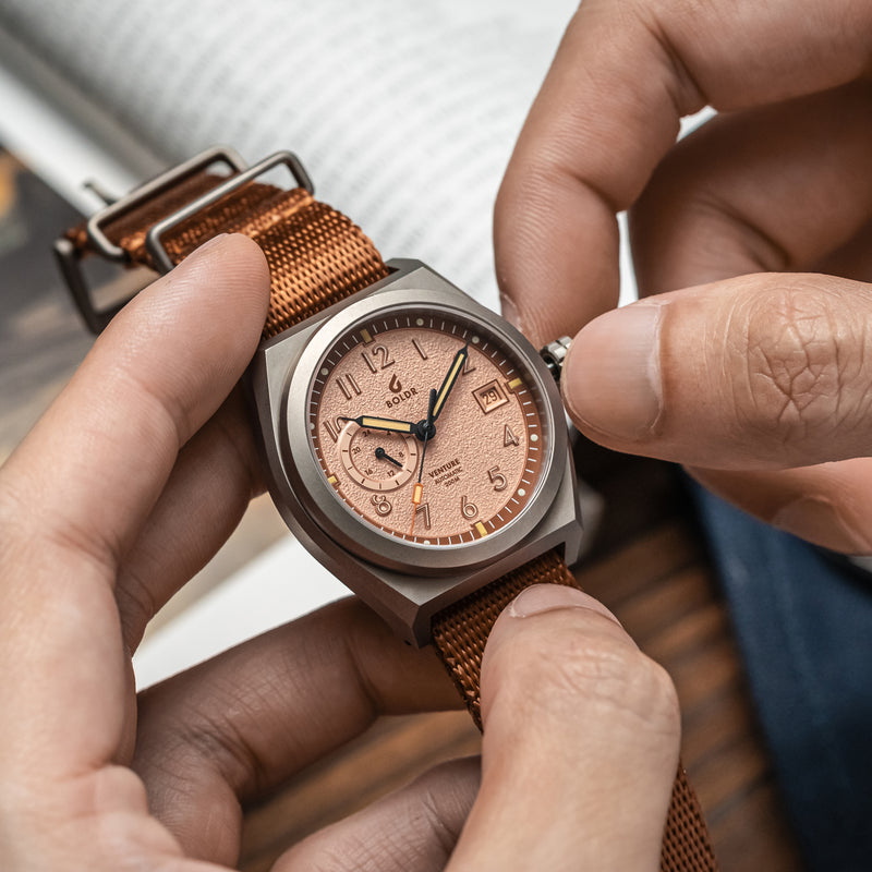 A close-up wrist shot of the BOLDR Venture Wayfarer, featuring a brushed titanium case and a textured copper dial. The watch is paired with a brown nylon strap, and the hand is wearing a grey knit sweater, adding a cozy yet rugged aesthetic.