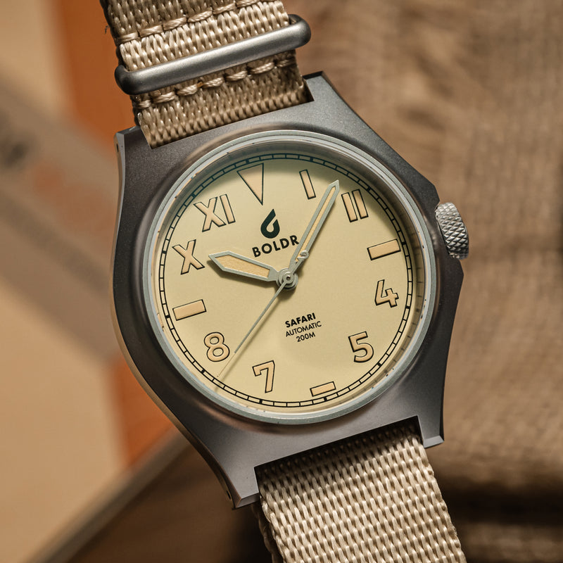 Close-up of a BOLDR watch with a beige dial and matching beige fabric strap, placed on a wooden surface. The watch face displays a mix of Roman and Arabic numerals, with the brand name ‘BOLDR’ and ‘Safari Automatic 200M’ text prominently visible. The watch is neatly arranged, showcasing its design and craftsmanship.