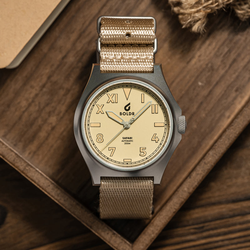 Close-up of a BOLDR watch with a beige dial and matching beige fabric strap, placed on a wooden surface. The watch face displays a mix of Roman and Arabic numerals, with the brand name ‘BOLDR’ and ‘Safari Automatic 200M’ text prominently visible. The watch is neatly arranged, showcasing its design and craftsmanship.