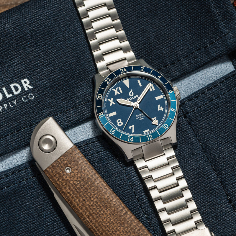 Close-up of a BOLDR Safari watch with a deep blue dial, Roman and Arabic numerals, and a stainless steel bracelet.
