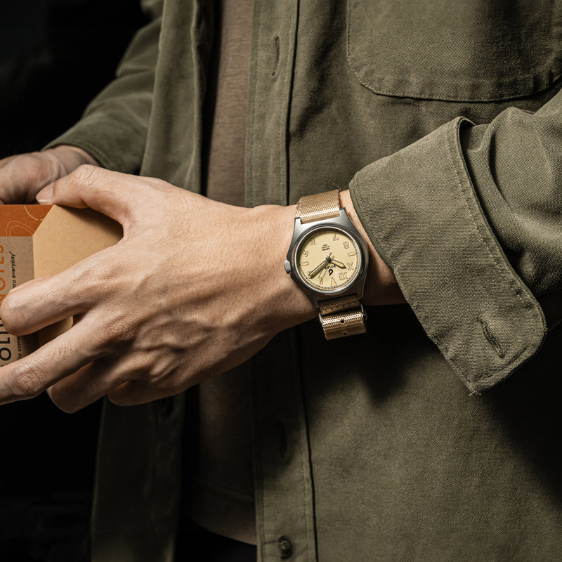 Close-up of a person’s wrist wearing a BOLDR watch with a beige dial and matching beige fabric strap. The watch face features large, bold numerals and simple, elegant hands. The person is dressed in a dark green jacket and dark pants, with the sleeve partially covering the watch.