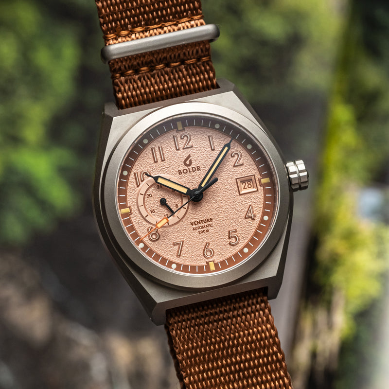 A close-up wrist shot of the BOLDR Venture Wayfarer, featuring a brushed titanium case and a textured copper dial. The watch is paired with a brown nylon strap.