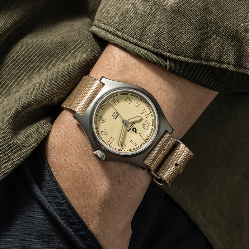 Close-up of a person’s wrist wearing a BOLDR watch with a beige dial and matching beige fabric strap. The watch face features large, bold numerals and simple, elegant hands. The person is dressed in a dark green jacket and dark pants, with the sleeve partially covering the watch.