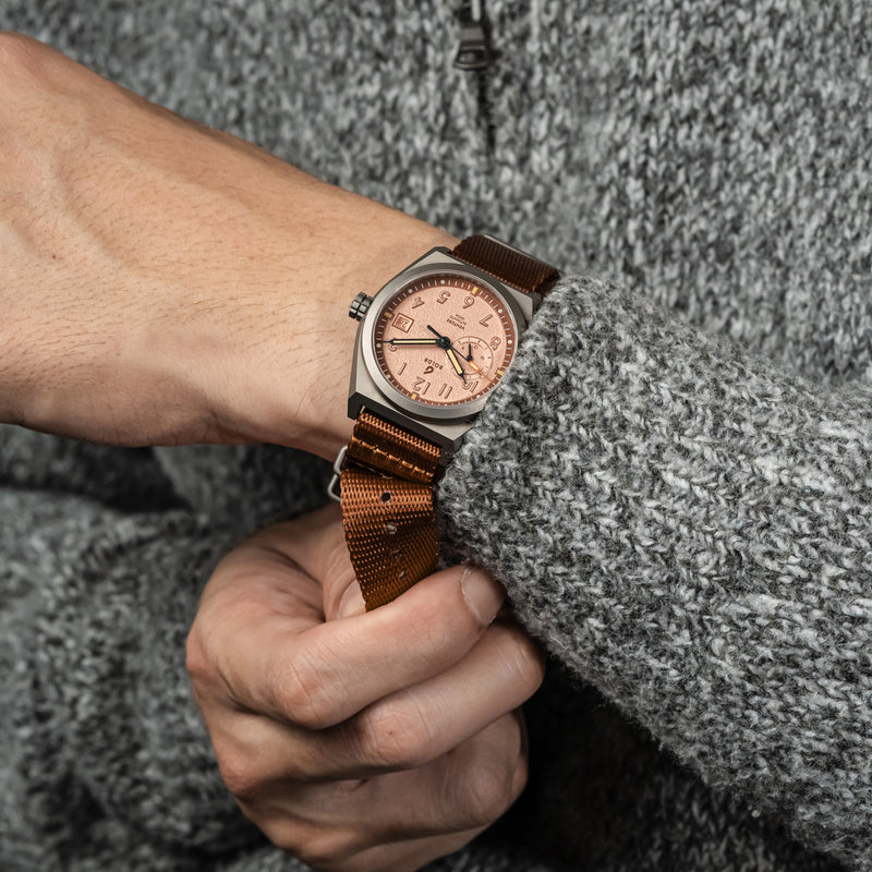 A close-up wrist shot of the BOLDR Venture Wayfarer, featuring a brushed titanium case and a textured copper dial. The watch is paired with a brown nylon strap, and the hand is wearing a grey knit sweater, adding a cozy yet rugged aesthetic.