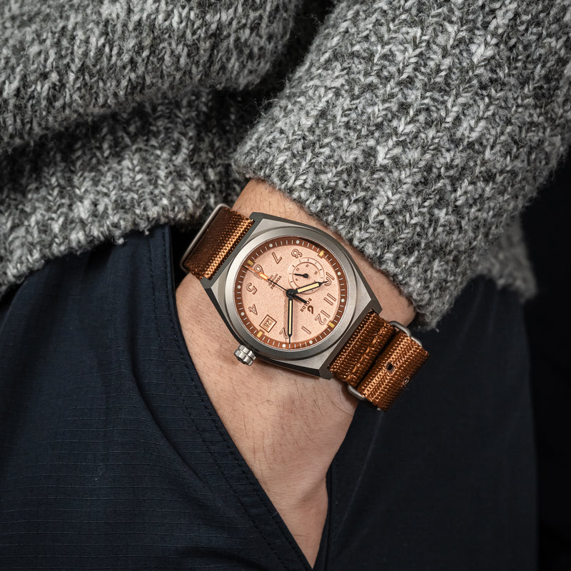 A close-up wrist shot of the BOLDR Venture Wayfarer, featuring a brushed titanium case and a textured copper dial. The watch is paired with a brown nylon strap, and the hand is wearing a grey knit sweater, adding a cozy yet rugged aesthetic.