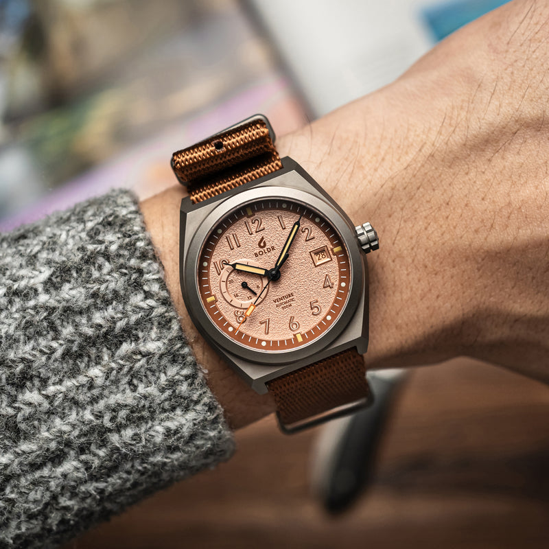 A close-up wrist shot of the BOLDR Venture Wayfarer, featuring a brushed titanium case and a textured copper dial. The watch is paired with a brown nylon strap, and the hand is wearing a grey knit sweater, adding a cozy yet rugged aesthetic.