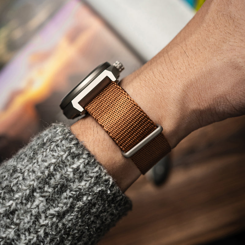 A close-up wrist shot of the BOLDR Venture Wayfarer, featuring a brushed titanium case and a textured copper dial. The watch is paired with a brown nylon strap, and the hand is wearing a grey knit sweater, adding a cozy yet rugged aesthetic.